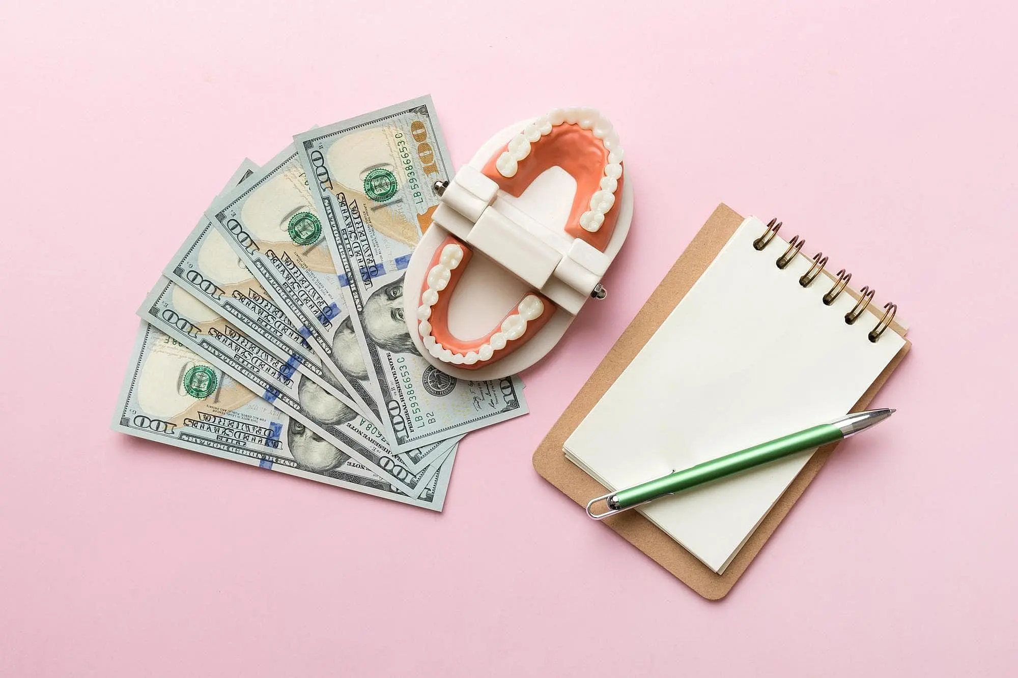 A dental model with metal braces, several $100 bills, a notepad, and a green pen are displayed on a pink surface at Happy Smiles Orthodontics in St. Augustine, FL.