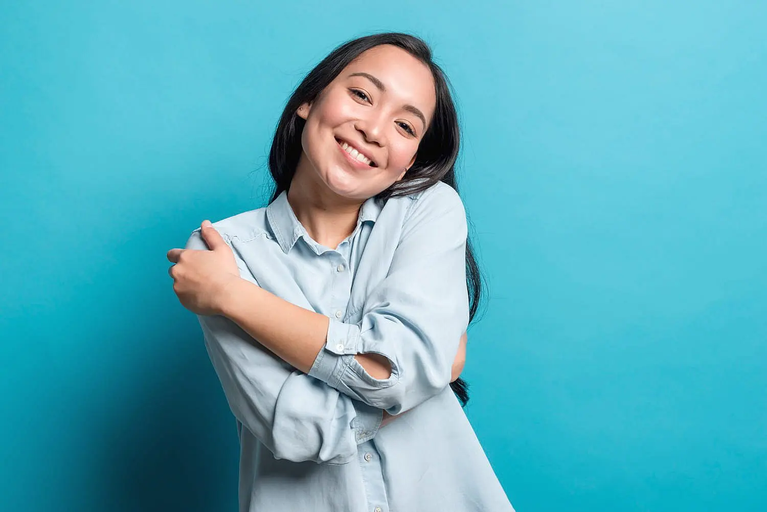 At Happy Smiles Orthodontics in St. Augustine, FL, a person with long dark hair and metal braces smiles confidently, hugging themselves against a plain blue background.