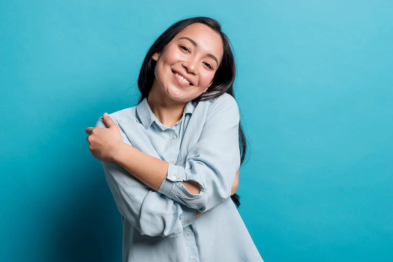 Confidently smiling with Invisalign aligners, a woman with long dark hair in a light blue shirt hugs herself against a blue background—showcasing the radiant results of Happy Smiles Orthodontics in St. Augustine, FL.