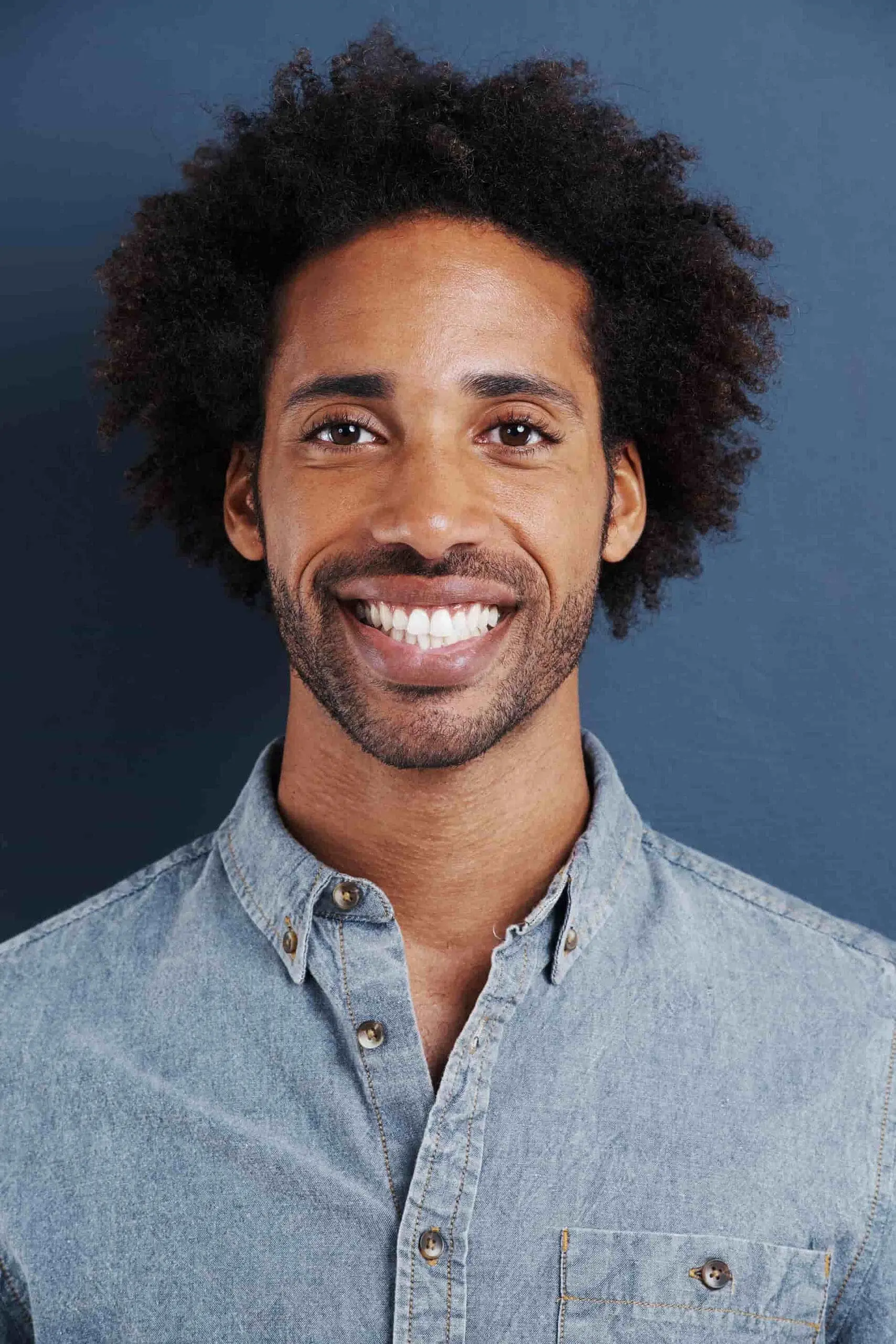 At Happy Smiles Orthodontics in St. Augustine, FL, a man with curly hair and a beard smiles confidently at the camera in a light denim shirt against a dark blue background, reflecting the approachable professionalism of an experienced orthodontist.
