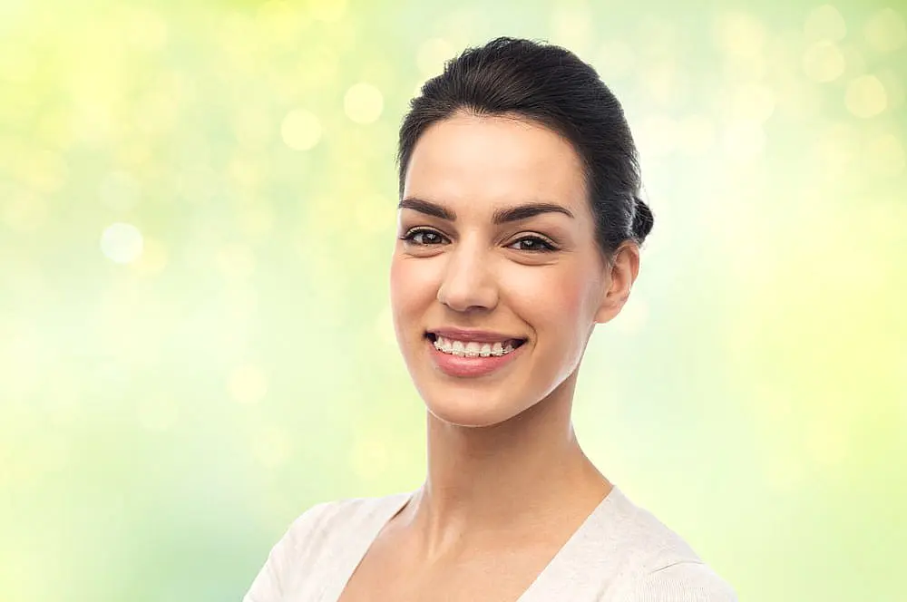 At Happy Smiles Orthodontics in St. Augustine, FL, a dark-haired woman smiles confidently at the camera against a blurred green and yellow backdrop, highlighting the results of adult orthodontics and clear braces.