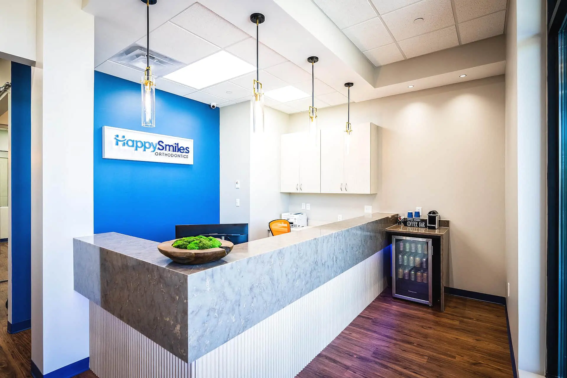 The reception area at Happy Smiles Orthodontics in St. Augustine, FL features a marble counter, blue accent wall, Happy Smiles sign, pendant lights, and a refreshment station for a warm welcome.