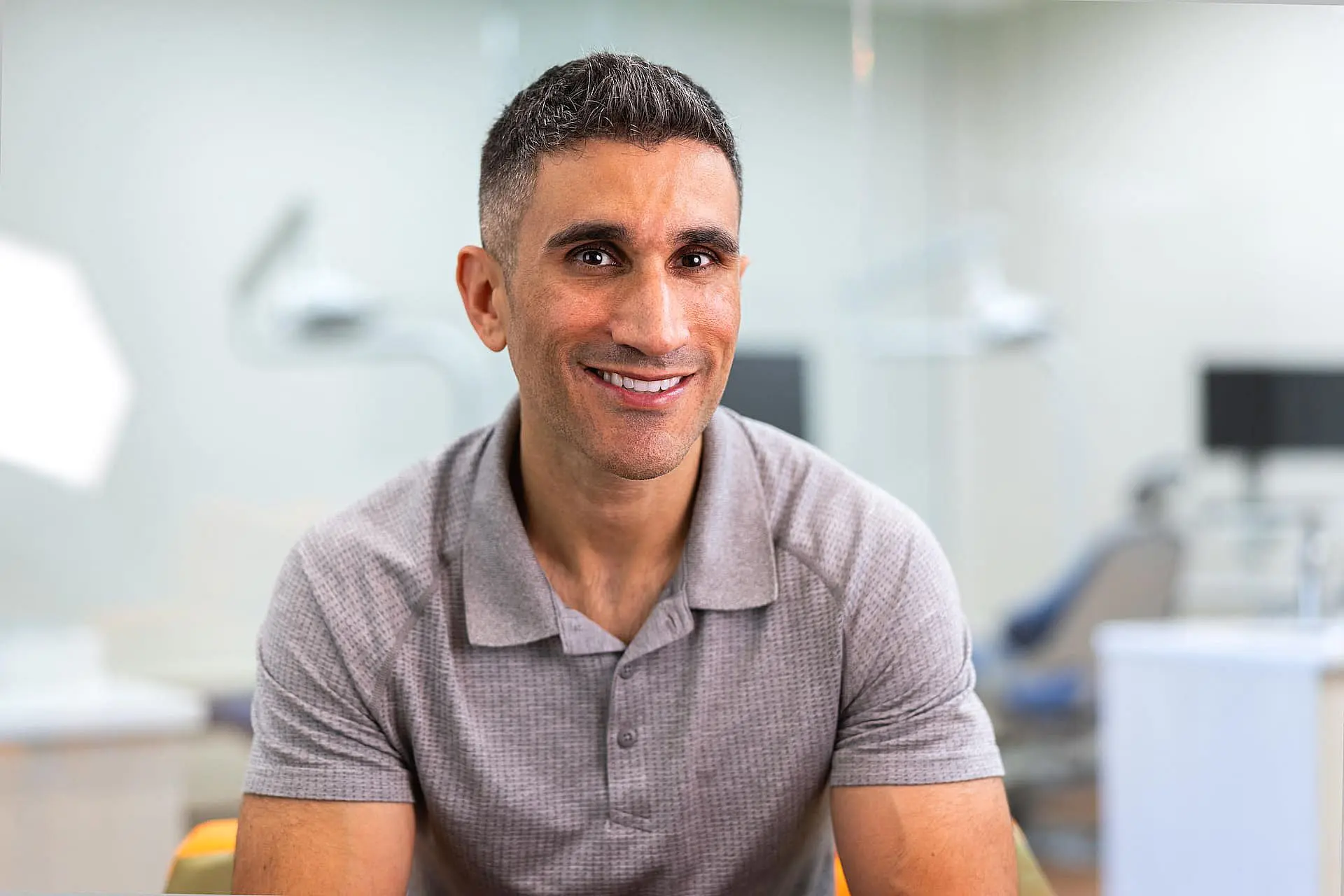 Orthodontics Expert Dr. Mark Komforti is pictured at Happy Smiles Orthodontics in St. Augustine, FL. With short dark hair and a grey polo shirt, he smiles at the camera in an indoor setting, with a softly blurred office or clinic background.