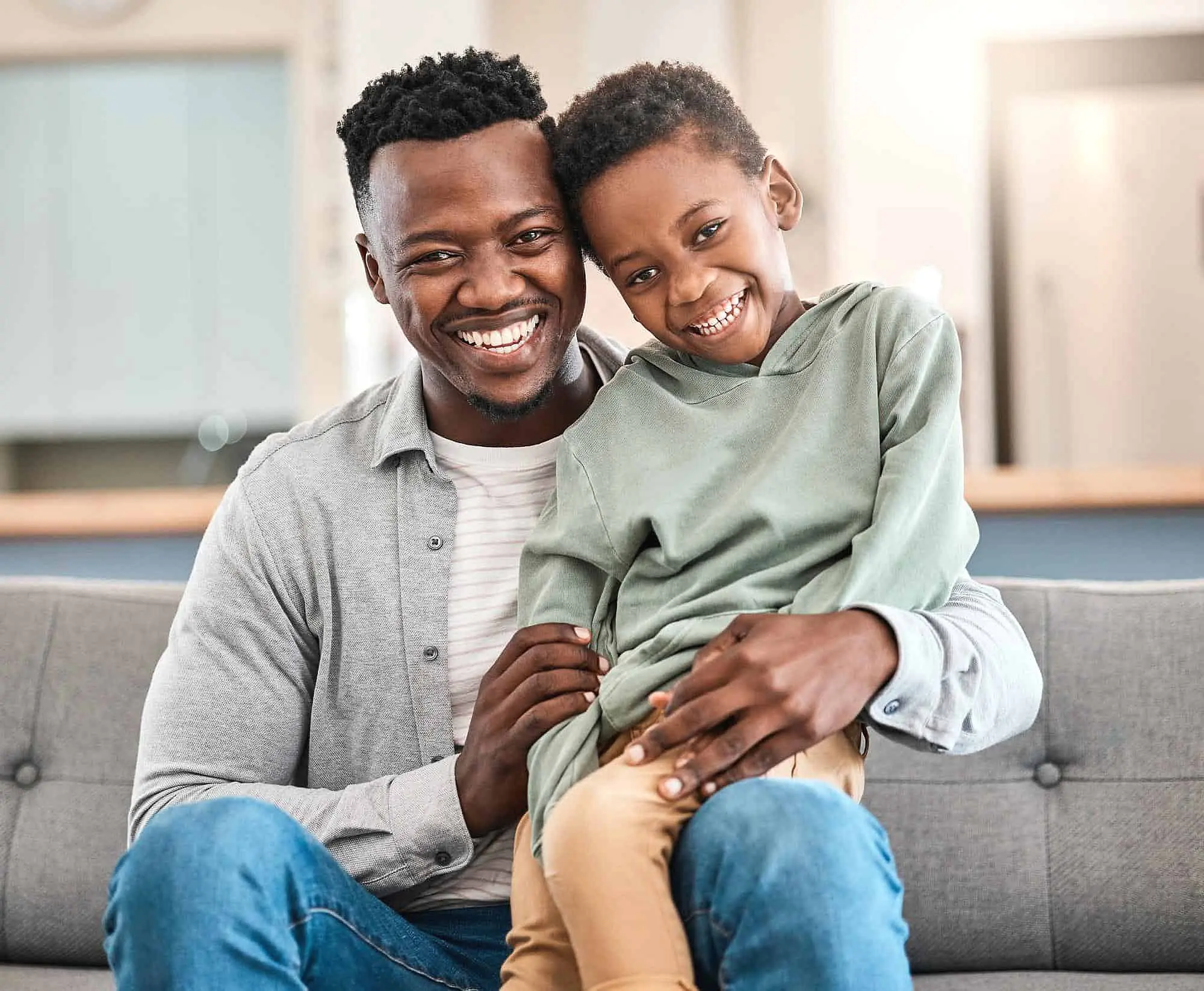 Both a man and a child sit together on a sofa, smiling confidently at the camera thanks to Happy Smiles Orthodontics in St. Augustine, FL.