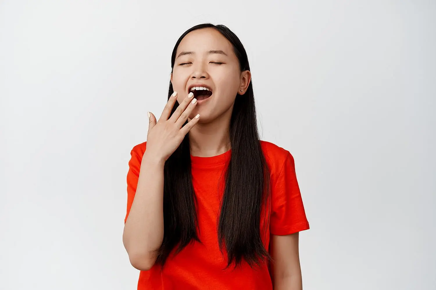 A young woman wearing a red t-shirt, eyes closed and hand near her mouth as she yawns, demonstrates an underbite for Happy Smiles Orthodontics in St. Augustine, FL, against a plain white background.