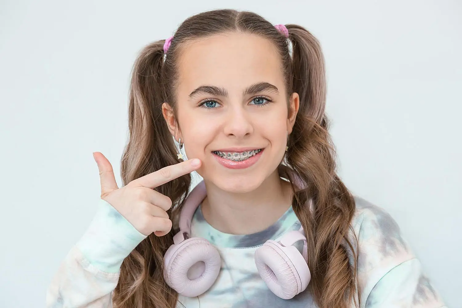 At Happy Smiles Orthodontics in St. Augustine, FL, a teen girl with long brown hair in pigtails, metal braces, a tie-dye shirt, and pink headphones around her neck smiles while pointing to her braces.