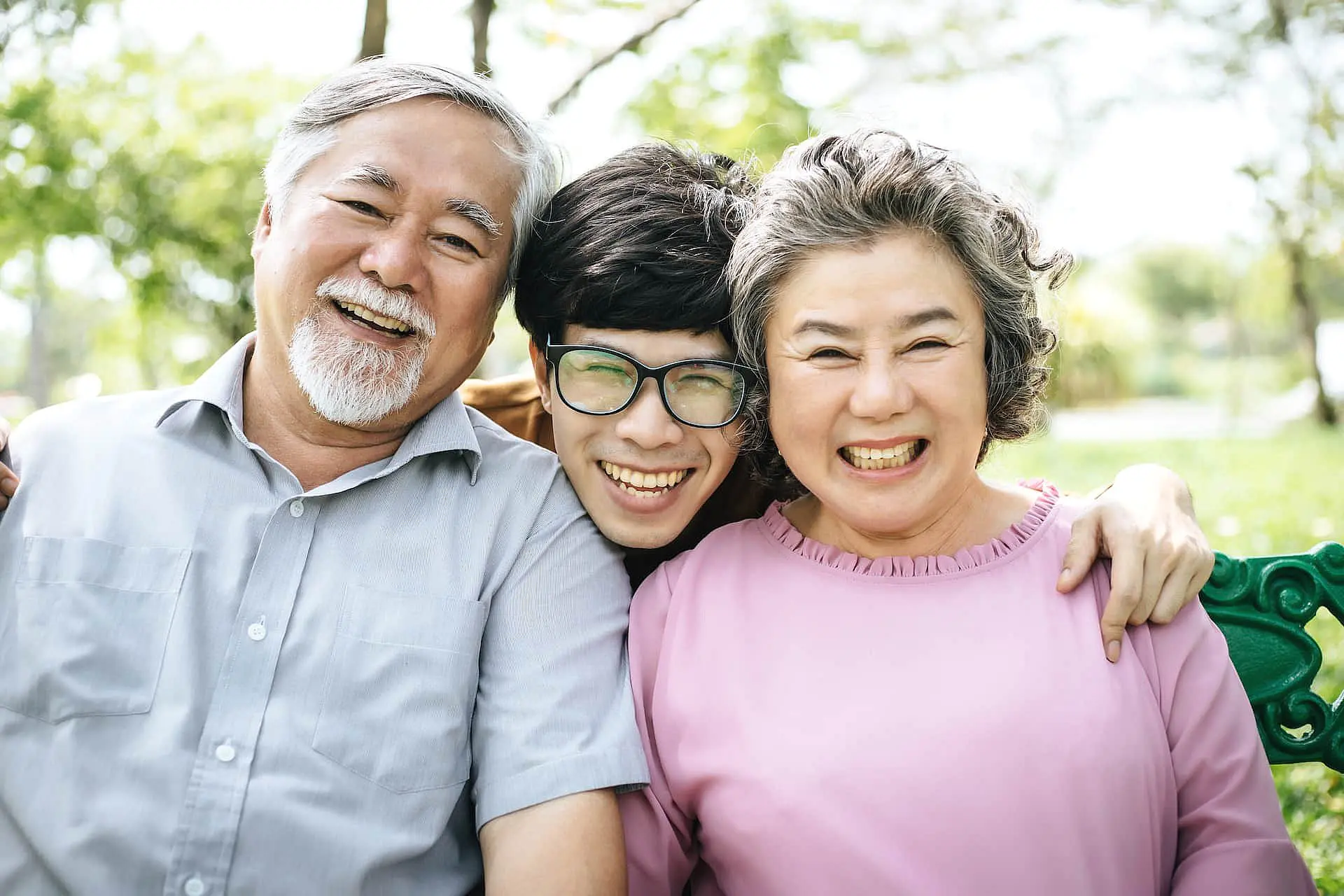 At Happy Smiles Orthodontics in St. Augustine, FL, three people sit closely on an outdoor bench, smiling at the camera as the younger person in the middle shows off their adult braces.