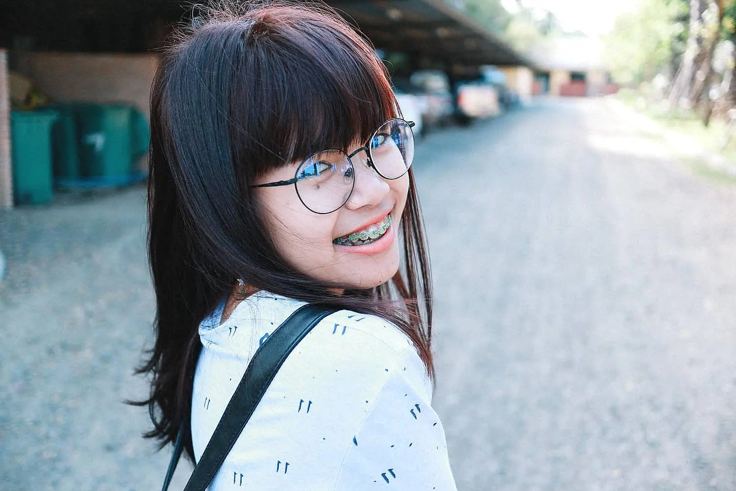 At Happy Smiles Orthodontics in St. Augustine, FL, a young person with glasses and braces demonstrates the advantages of adult orthodontics, smiling confidently over their shoulder on a gravel path lined with trees and parked cars.