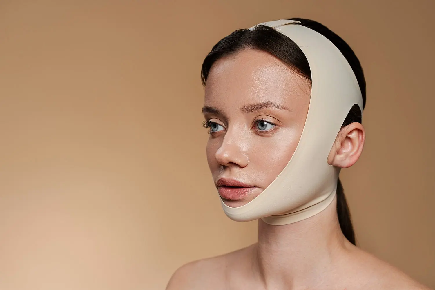 At Happy Smiles Orthodontics in St. Augustine, FL, a woman with brown hair is shown against a neutral background wearing a beige facial compression bandage around her chin and head, typically used following jaw surgery.