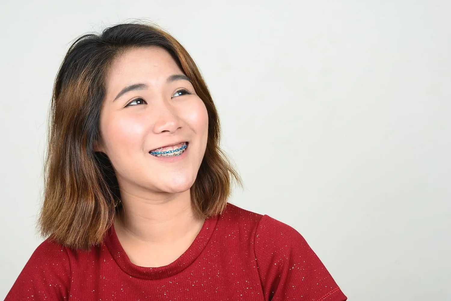 A young woman with shoulder-length brown hair, wearing a red shirt and adult braces, smiles and looks slightly left against a plain light background—representing Happy Smiles Orthodontics in St. Augustine, FL.