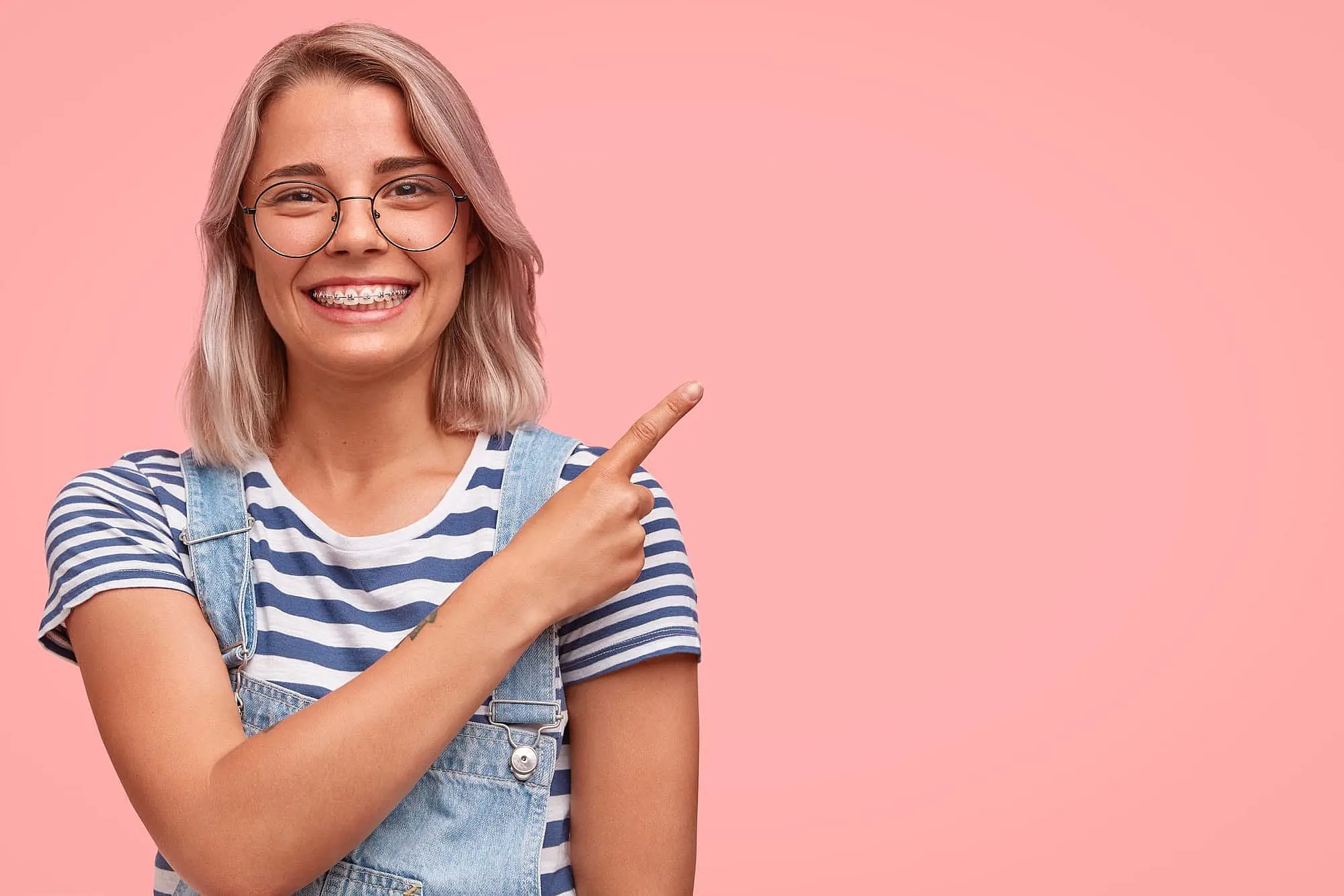 Showcasing the benefits of adult orthodontics, a young person with glasses and braces smiles and points to the right against a plain pink background—representing Happy Smiles Orthodontics in St. Augustine, FL.