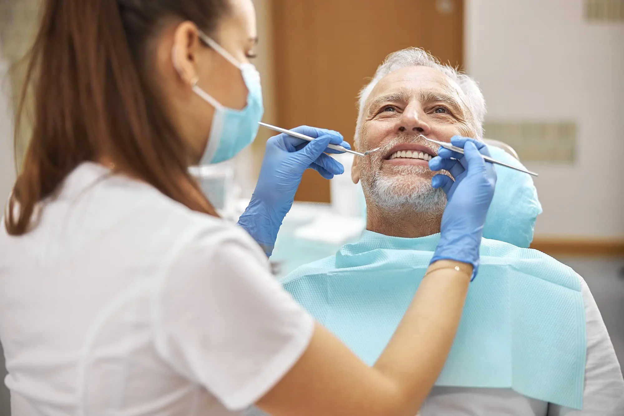 At Happy Smiles Orthodontics in St. Augustine, FL, a dental professional in gloves and a mask examines an older man’s teeth as he smiles in the dental chair, likely discussing jaw surgery options for better oral health.