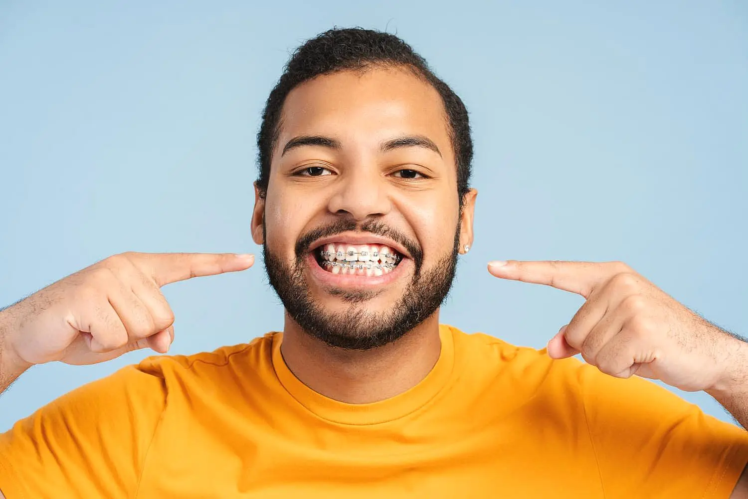 A man in a yellow shirt smiles and points at his adult braces against a plain blue background, representing Happy Smiles Orthodontics in St. Augustine, FL.