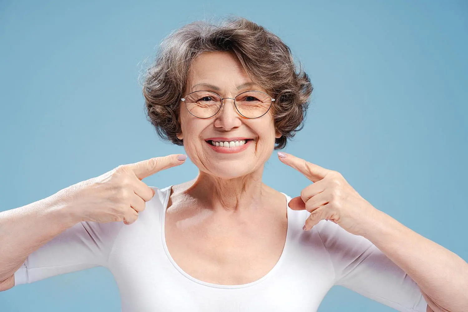 A smiling older woman with short gray hair and glasses points to her teeth against a plain light blue background, highlighting the benefits of Orthodontic Insurance Plans at Happy Smiles Orthodontics in St. Augustine, FL.