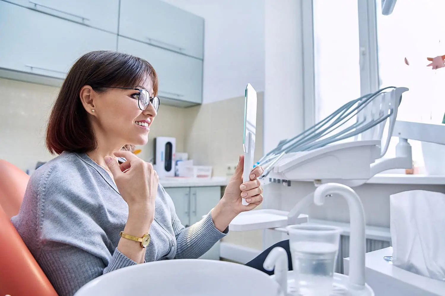 At Happy Smiles Orthodontics in St. Augustine, FL, a woman sits in a dental chair and confidently smiles at her reflection in a hand mirror after completing her adult orthodontic treatment.
