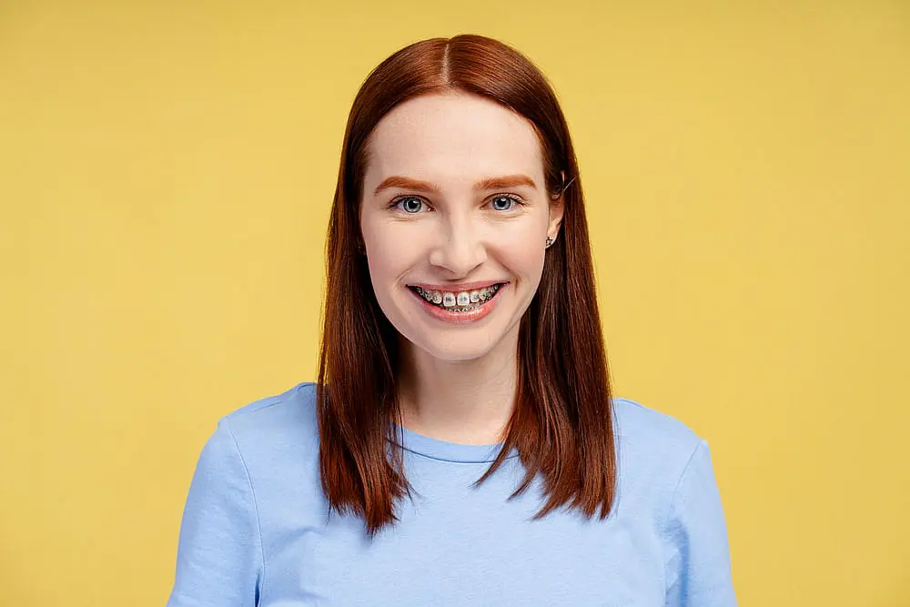 At Happy Smiles Orthodontics in St. Augustine, FL, a young woman with straight red hair and traditional metal braces smiles at the camera, dressed in a light blue shirt against a yellow background.