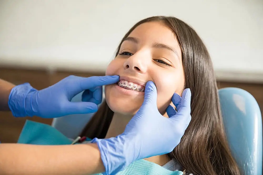 Happy Smiles Orthodontics At Happy Smiles Orthodontics in St. Augustine, FL, a dentist wearing blue gloves checks a smiling girl's braces and discusses the cost with her during a dental visit.