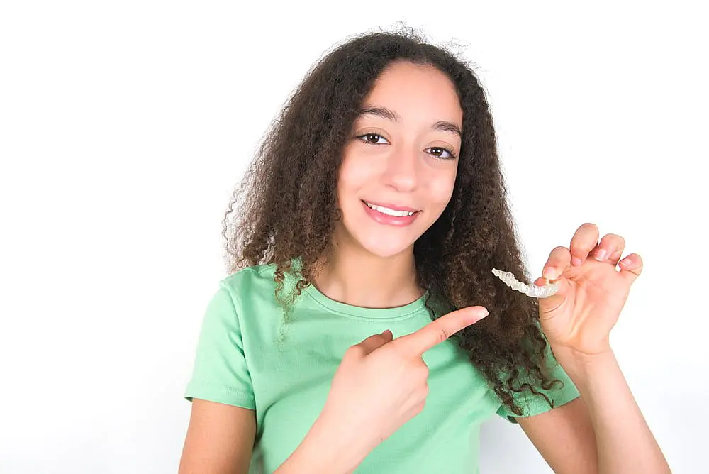 A girl with curly hair wearing a green shirt smiles and points to Invisalign® Clear Aligners she holds, highlighting Happy Smiles Orthodontics in St. Augustine, FL.