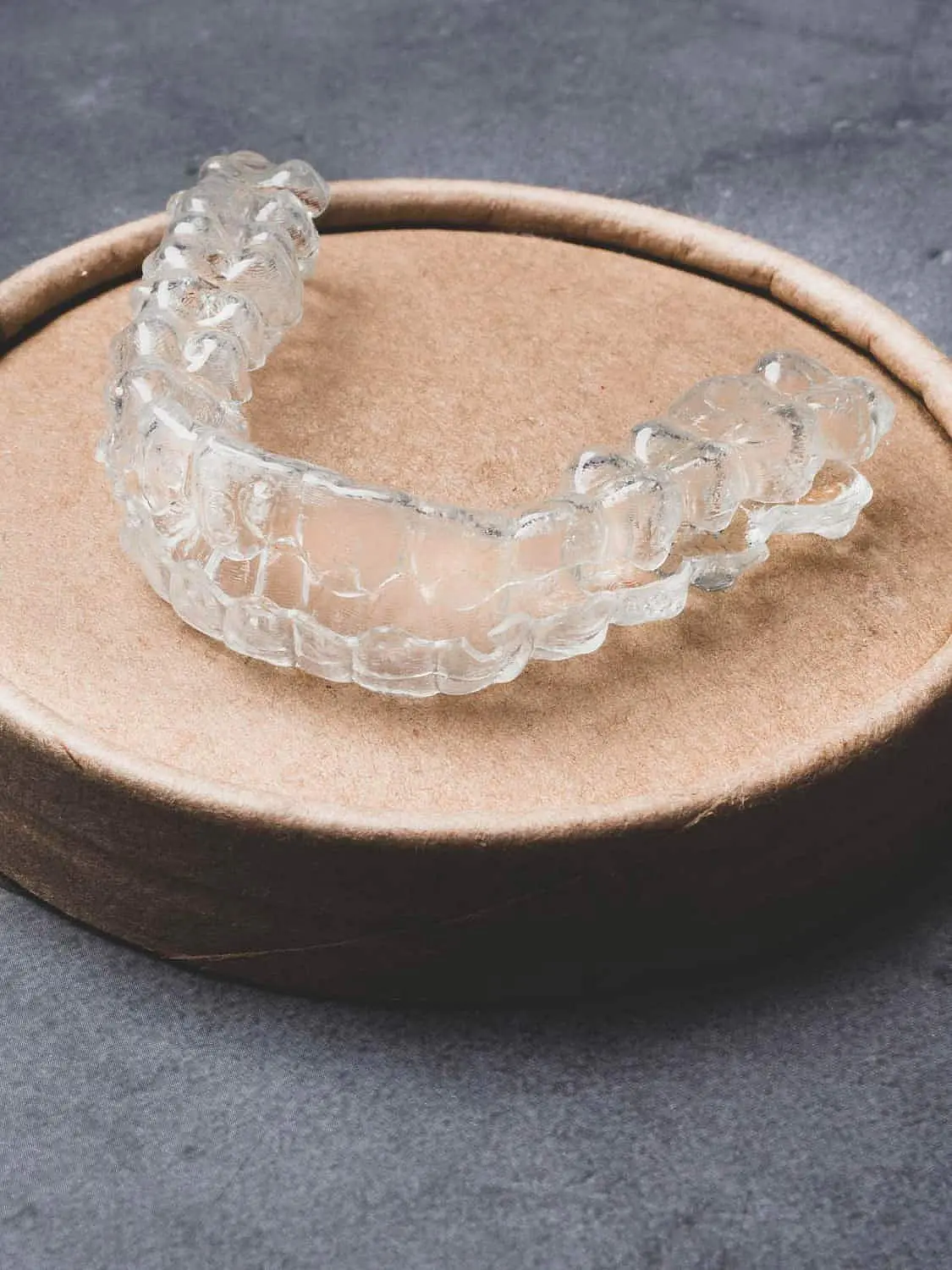 Showcasing an easy Invisalign payment plan, a clear dental aligner sits on a round cardboard container against a gray textured surface at Happy Smiles Orthodontics in St. Augustine, FL.