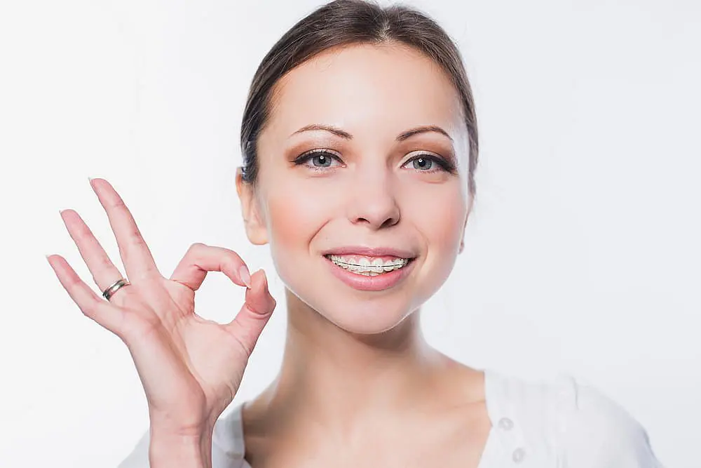 A woman with clear braces flashes an "OK" hand gesture and smiles, representing Happy Smiles Orthodontics in St. Augustine, FL, against a plain white background.