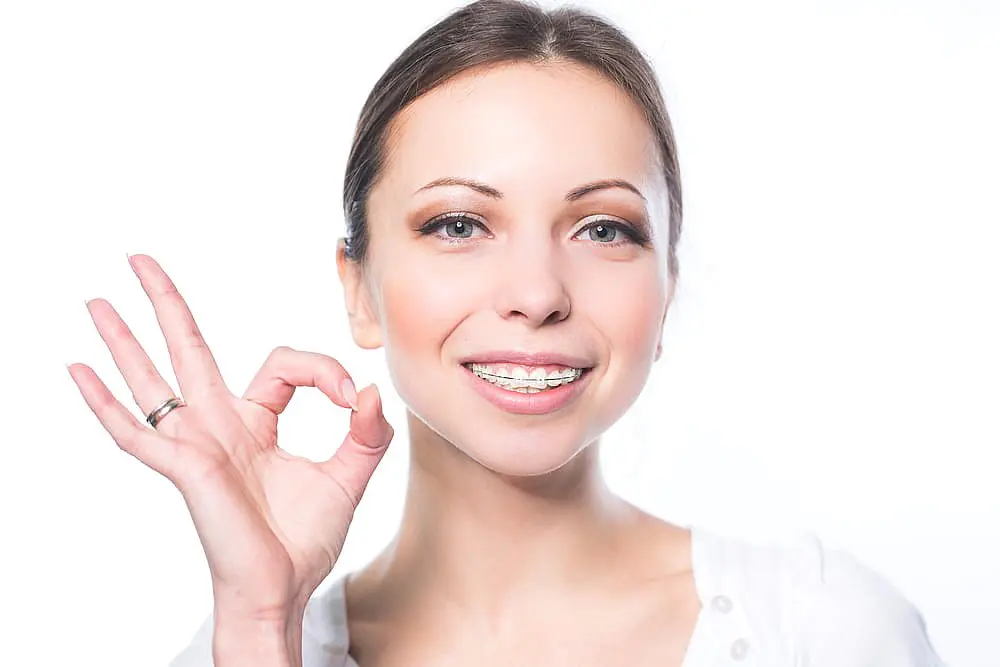 A young woman with clear ceramic braces smiles and makes an "OK" hand gesture against a white background at Happy Smiles Orthodontics in St. Augustine, FL.