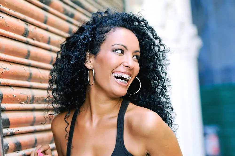 A woman with curly hair and braces, wearing hoop earrings and a black tank top, smiles while standing by a rusty metal shutter—highlighting adult orthodontics at Happy Smiles Orthodontics in St. Augustine, FL.