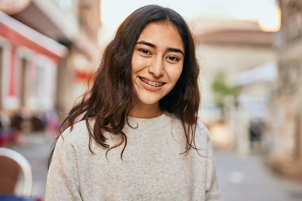 Happy Smiles Orthodontics Smiling confidently, a young woman with long brown hair and braces poses outdoors on a city street after discovering affordable braces cost at Happy Smiles Orthodontics in St. Augustine, FL.