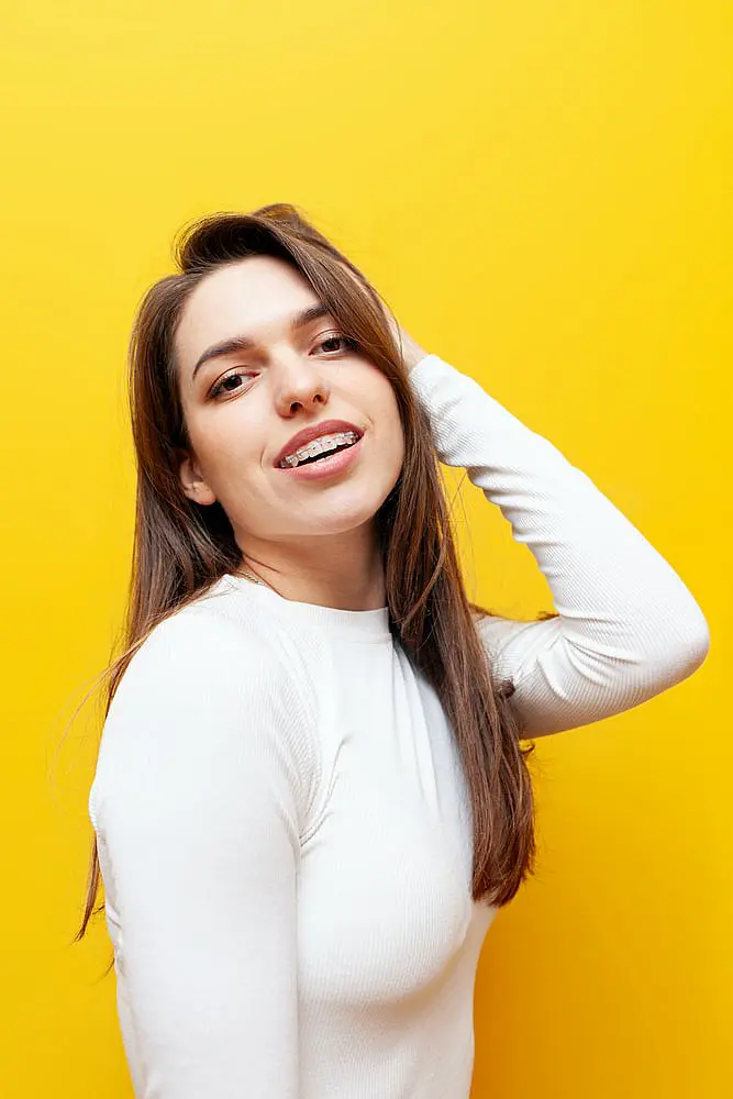 A young woman with long brown hair, clear ceramic braces, and a white long-sleeve shirt smiles against a bright yellow background at Happy Smiles Orthodontics in St. Augustine, FL.