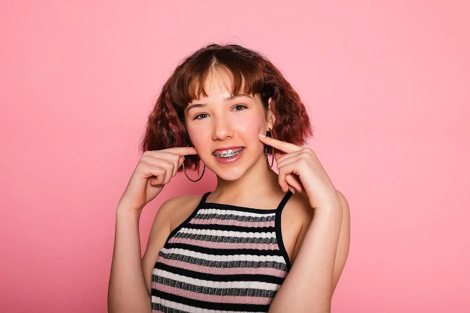 A teen girl with short brown hair and braces stands in front of a solid pink background, wearing a striped sleeveless top. She smiles and points to her cheeks, highlighting her Invisalign vs Braces experience at Happy Smiles Orthodontics in St. Augustine, FL.