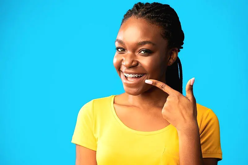 A young woman in a yellow shirt smiles and points to her braces, highlighting the benefits of adult orthodontics at Happy Smiles Orthodontics in St. Augustine, FL, set against a vibrant blue backdrop.