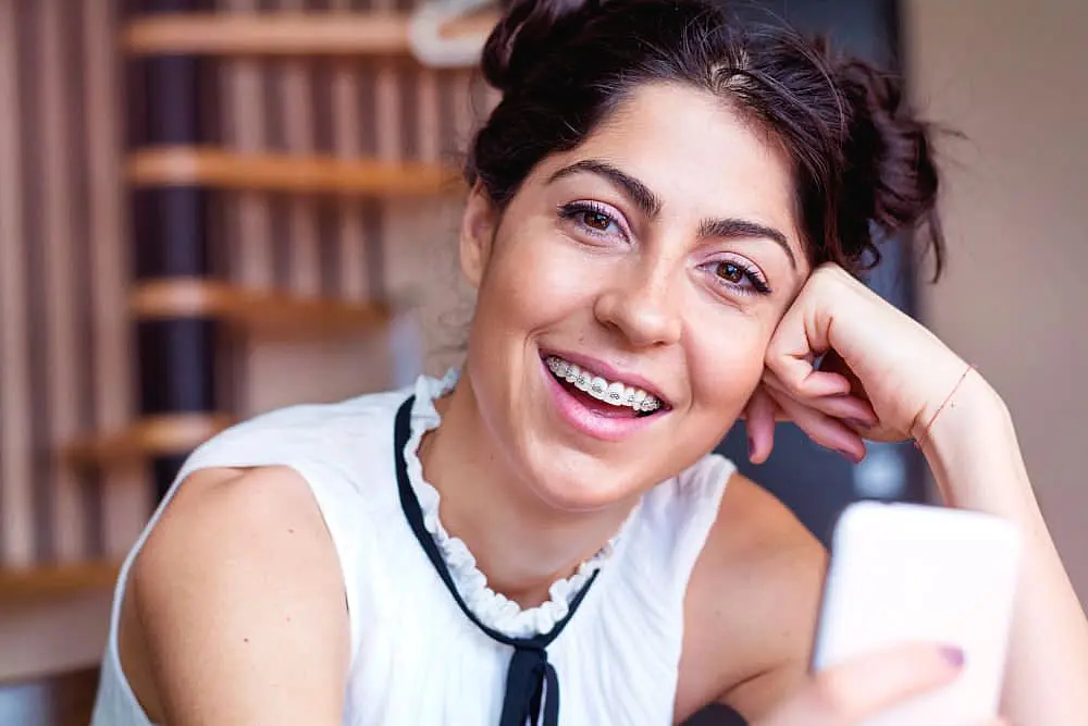 At Happy Smiles Orthodontics in St. Augustine, FL, a young woman with dark hair in buns and braces from adult orthodontics smiles indoors, holding a smartphone and wearing a white sleeveless top.