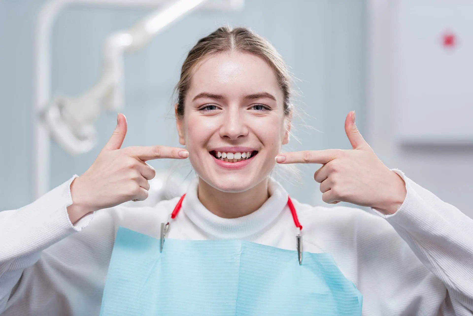 At Happy Smiles Orthodontics in St. Augustine, FL, a young woman wearing a dental bib smiles and points to the gap in her front teeth, with dental equipment in the background—showcasing her overbite.