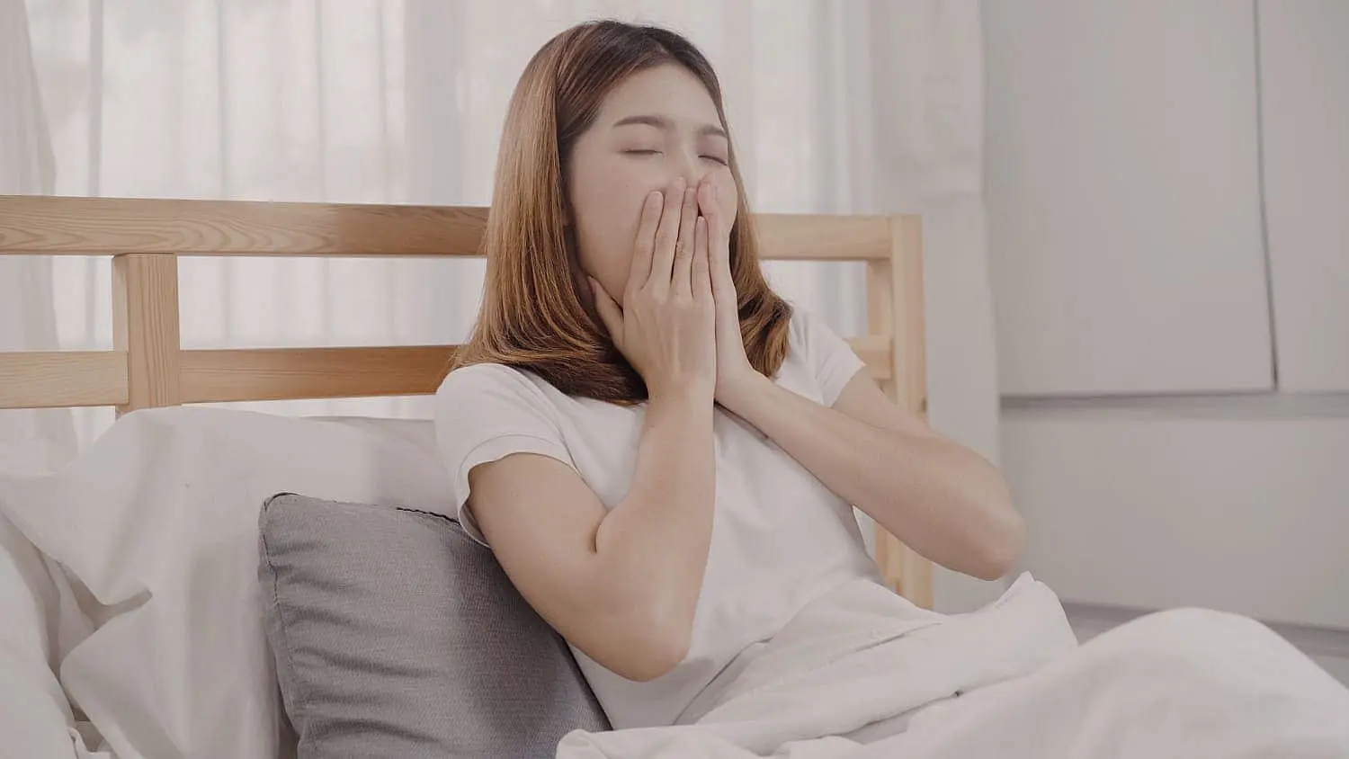 A woman sits up in bed, yawning and covering her mouth with her hands, suggesting tiredness or mouth breathing—an issue addressed by Happy Smiles Orthodontics in St. Augustine, FL.