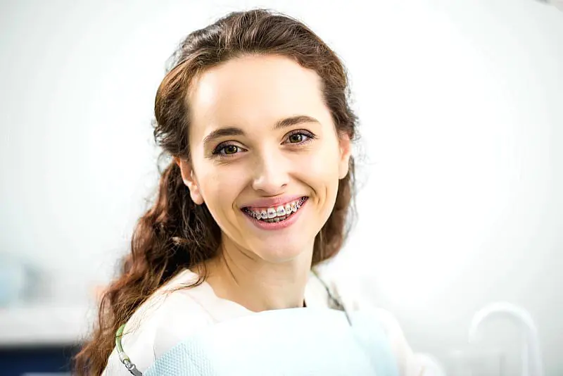 Smiling in a dental chair with metal braces and a dental bib, a young woman highlights the benefits of adult orthodontics at Happy Smiles Orthodontics in St. Augustine, FL.